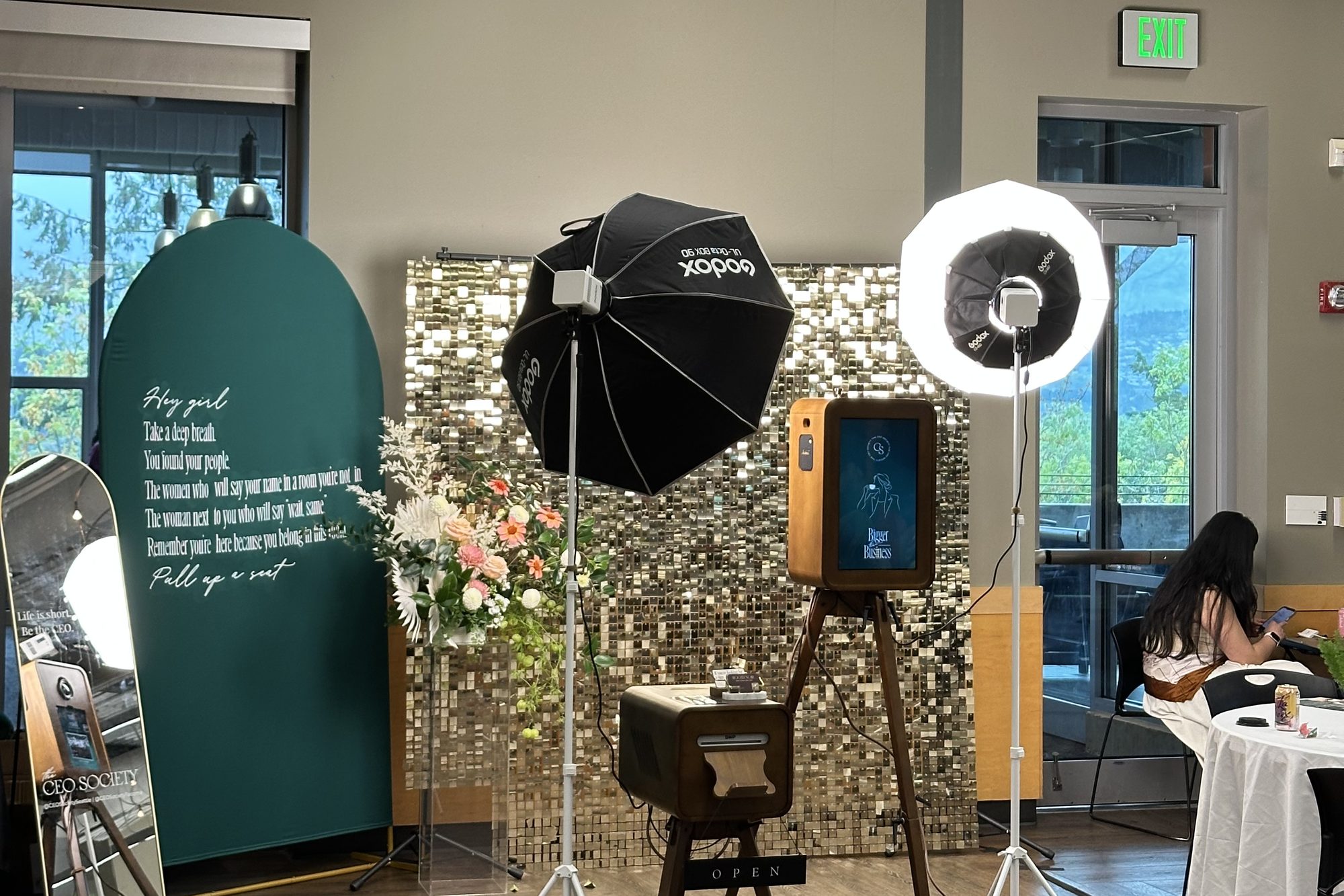The booth set up in a conference room with a sequin wall, a green arch, and flowers, tables in the background